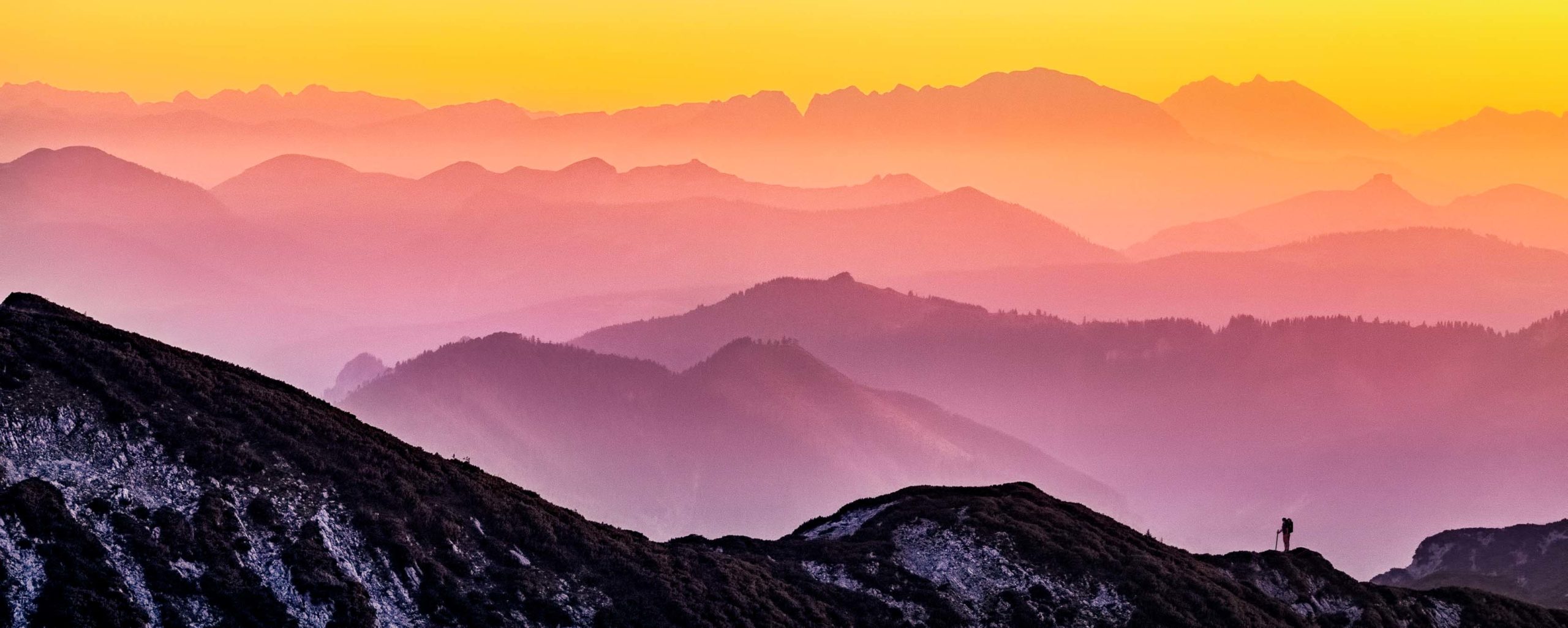 Hiker climbs ride in front of yellow and pink tinted mountains